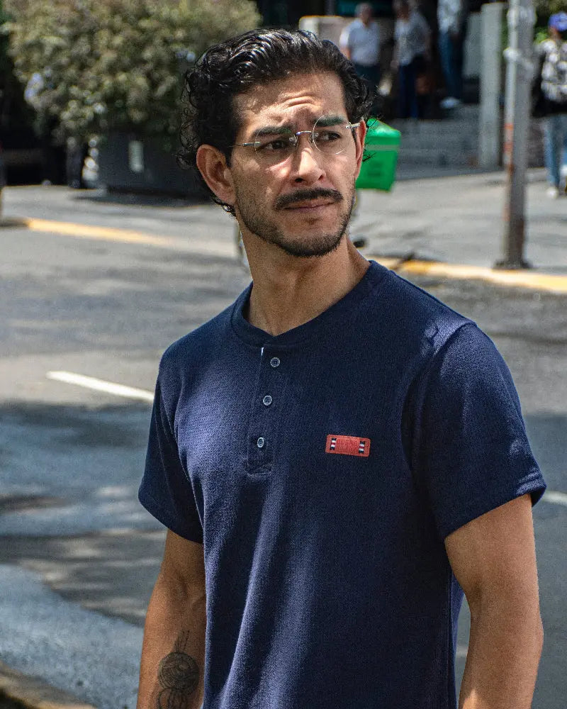 Hombre con gafas de sol y reloj de perfil en un parque, vistiendo una camiseta tipo Henley azul marino texturizada de la marca Dubrovnik con botones abiertos