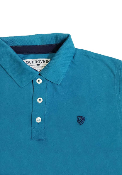 Primer plano del cuello de un polo infantil en color turquesa oscuro de la marca Dubrovnik, destacando el contraste azul marino en la tira interna del cuello, tres botones claros y el escudo bordado en azul marino sobre el pecho.