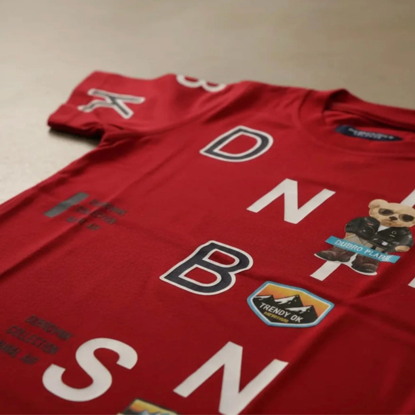 Primer plano en ángulo de camiseta roja para niño con parches de montaña, oso con gafas de sol y letras blancas en relieve