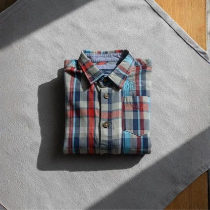 Camisa infantil de cuadros en tonos rojo, azul y gris perfectamente doblada sobre una superficie neutra con luz natural