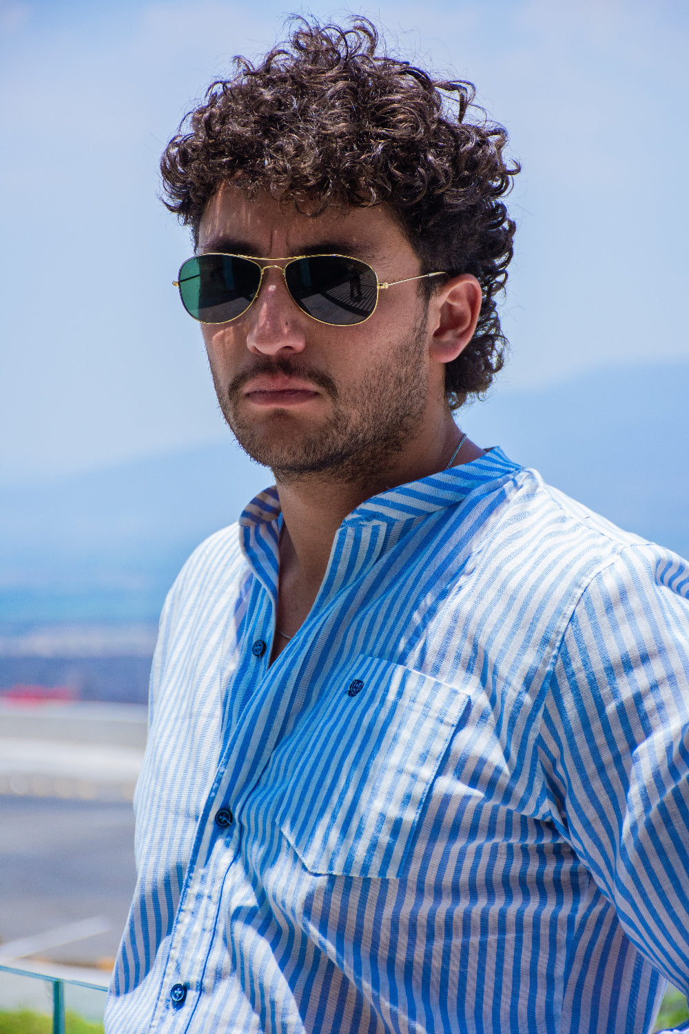 Modelo masculino con gafas de sol luciendo una camisa de lino a rayas azules y blancas con cuello mao y detalle de logo en el bolsillo