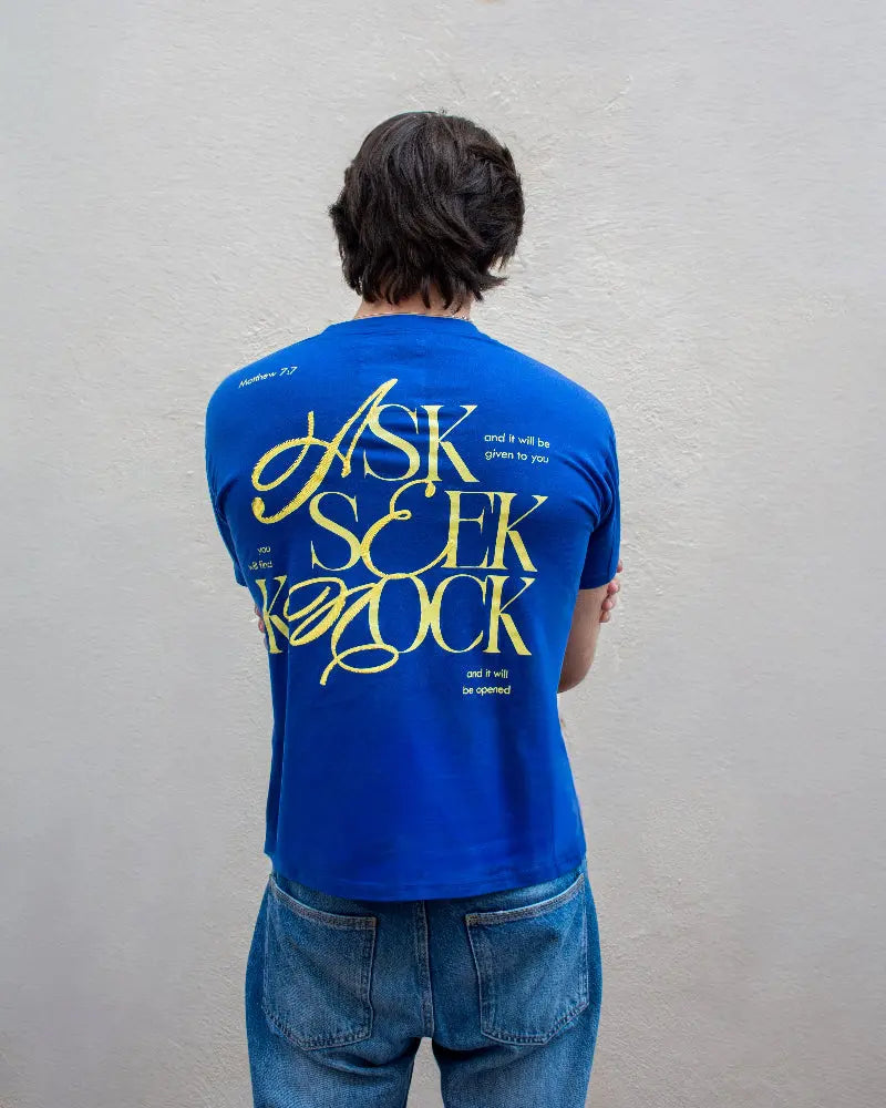 Vista trasera de hombre con camiseta azul destacando un gran diseño tipográfico amarillo con el texto "ASK SEEK KNOCK" y pasaje bíblico Matthew 7:7 sobre fondo blanco