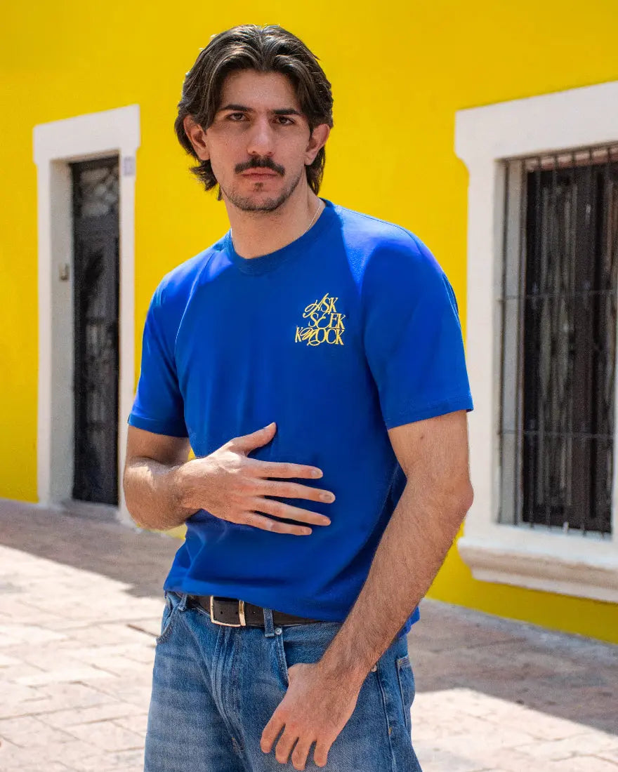 Hombre con bigote posando de frente con una camiseta azul royal de Dubrovnik, mostrando el logotipo amarillo "Ask Seek Knock" en el pecho sobre un fondo de pared amarilla.