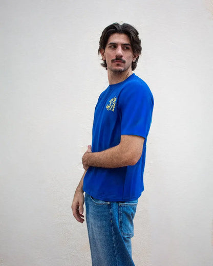 Hombre de perfil con bigote vistiendo camiseta azul royal con gráfico amarillo "Ask Seek Knock" en el pecho y pantalones vaqueros sobre fondo blanco