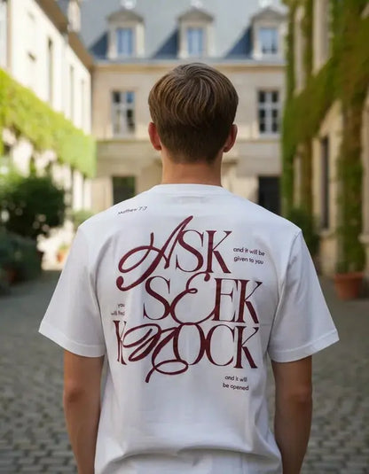 vista trasera de hombre con camiseta blanca destacando un diseño tipográfico en color guinda con el texto "ASK SEEK KNOCK" y referencia a Matthew 7:7 en un entorno urbano