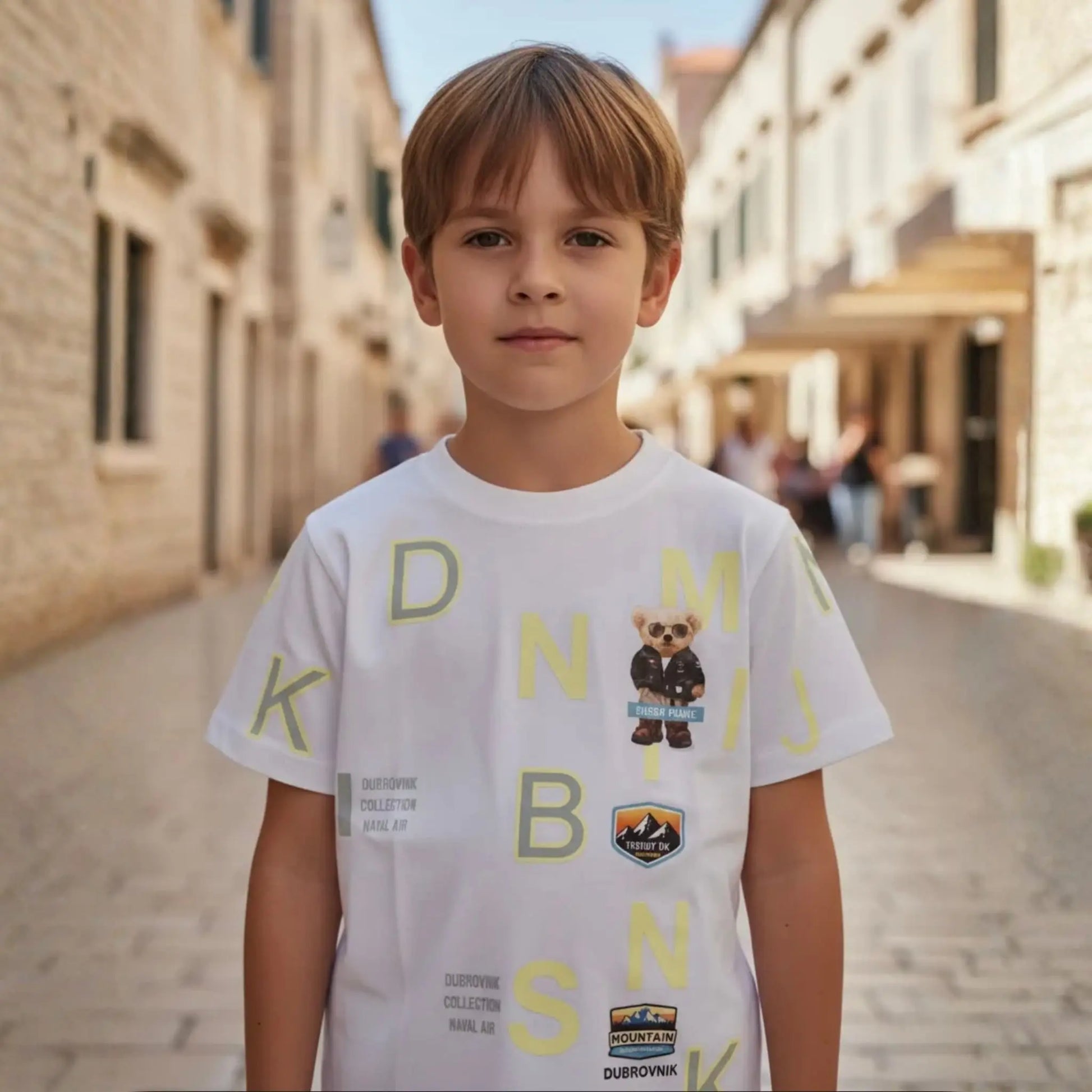 Niño con camiseta blanca de manga corta con gráficos amarillos y parche de oso en una calle antigua de piedra