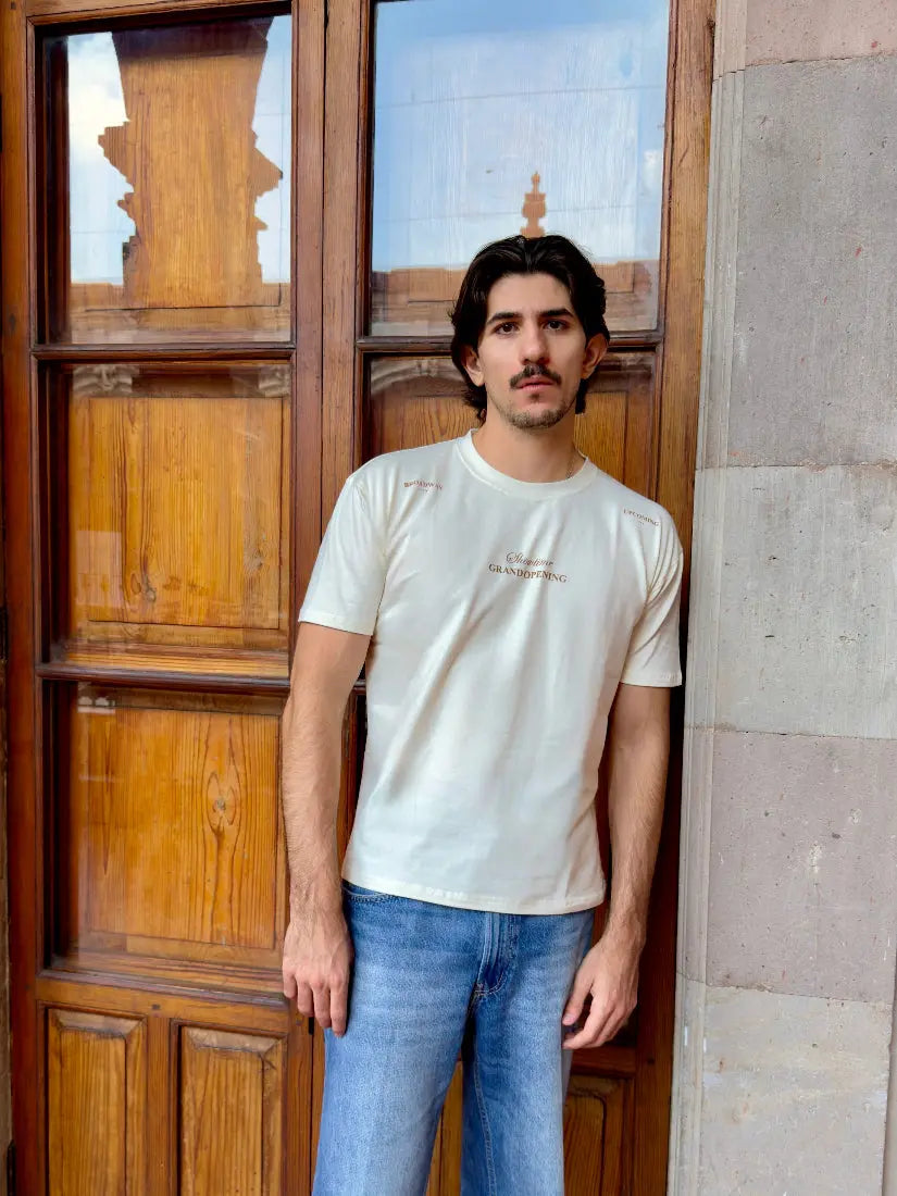 Hombre con bigote posando con camiseta color blanco hueso de la colección Showtime de Dubrovnik, con el texto "Grand Opening" en el pecho y detalles en los hombros, frente a una puerta de madera antigua
