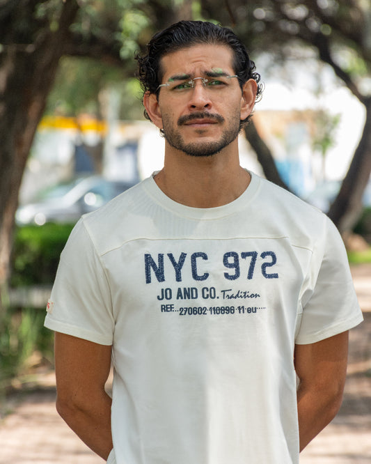 Hombre con gafas posando en un parque con camiseta blanca de manga corta con gráfico "NYC 972 JO AND CO Tradition