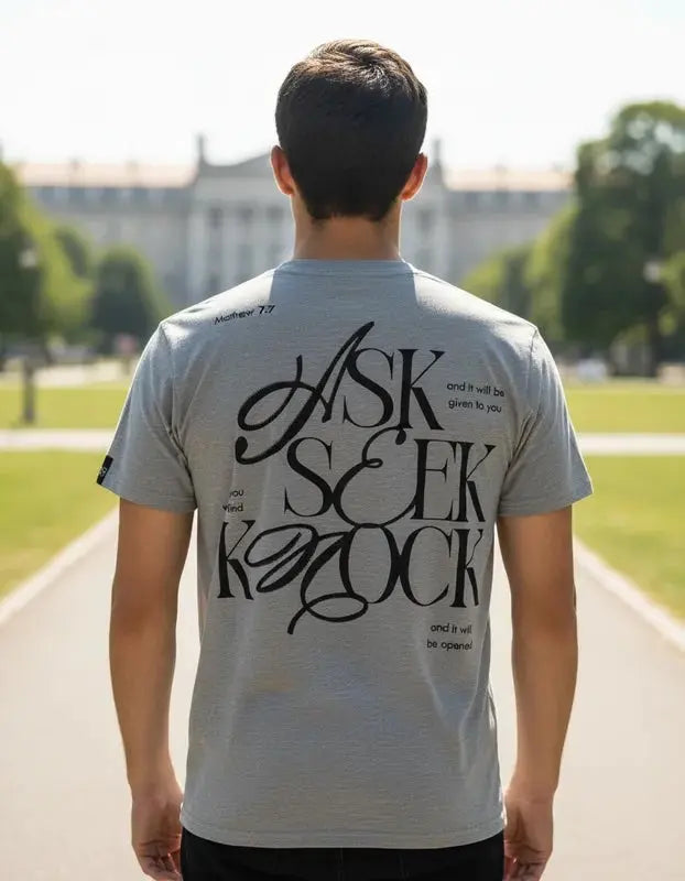 ista trasera de hombre con camiseta gris jaspeada mostrando el diseño tipográfico "ASK SEEK KNOCK" en color negro con referencia bíblica