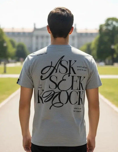 ista trasera de hombre con camiseta gris jaspeada mostrando el diseño tipográfico "ASK SEEK KNOCK" en color negro con referencia bíblica