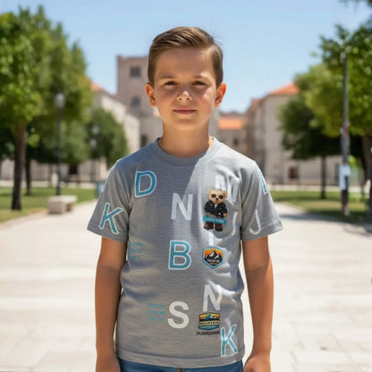 Niño posando con camiseta gris de manga corta con letras blancas y parches de montaña y oso en una plaza soleada.