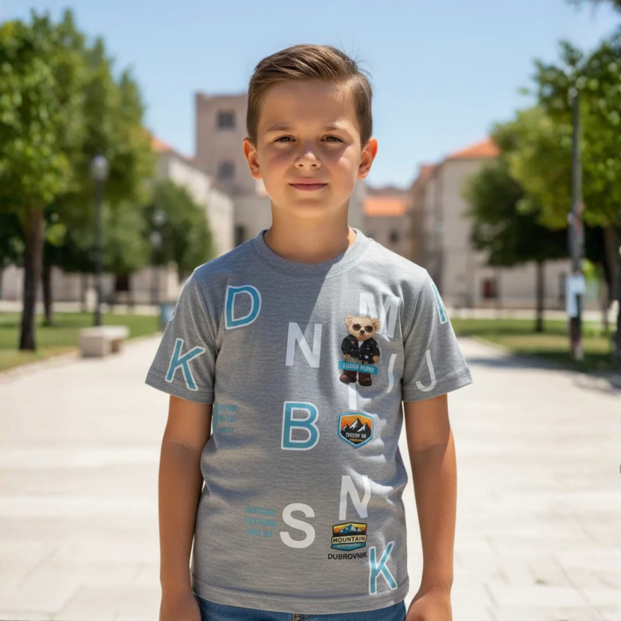 Niño posando con camiseta gris de manga corta marca Dubrovnik, con parches de oso y montaña en una plaza exterior soleada