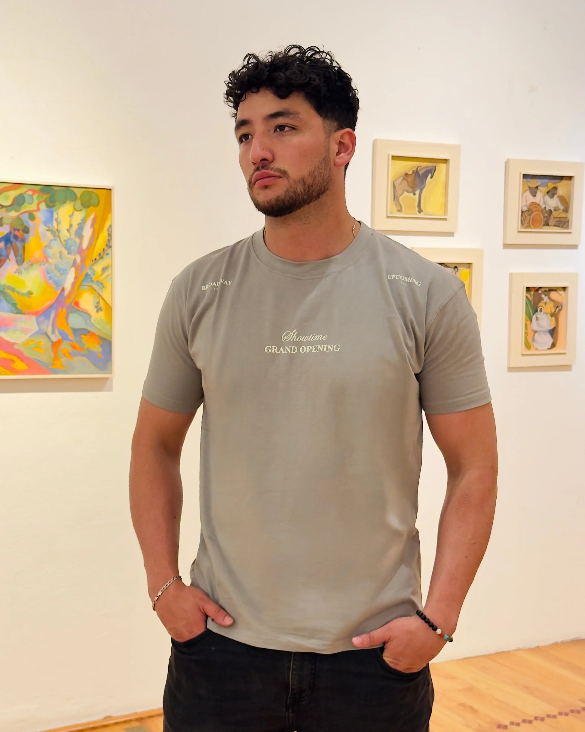 Hombre con pelo rizado posando con camiseta color gris piedra de la marca Dubrovnik, con el texto "Showtime Grand Opening" en el pecho y detalles en los hombros, ambientado en una galería de arte