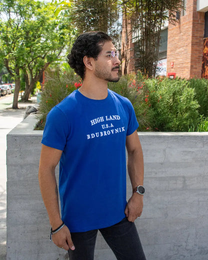 Hombre con gafas y bigote mirando hacia un lado, vistiendo camiseta Henley azul marino texturizada con botones y parche naranja.