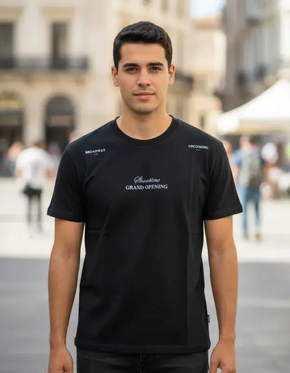 Hombre posando en exteriores con camiseta negra de la colección Showtime de Dubrovnik, con el texto "Grand Opening" en el pecho y detalles tipográficos blancos en los hombros