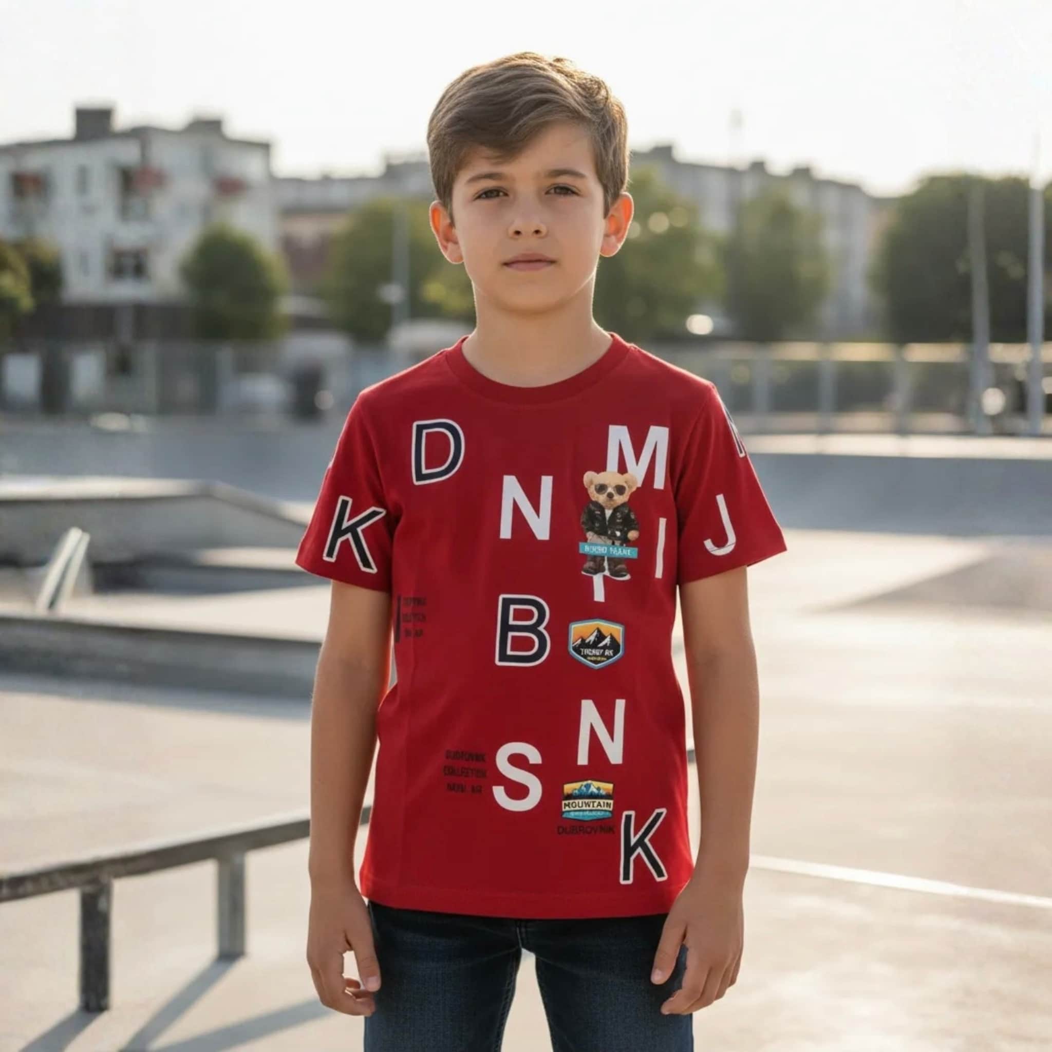 Niño posando con camiseta roja de manga corta con estampados blancos de letras y parches de oso en un entorno urbano