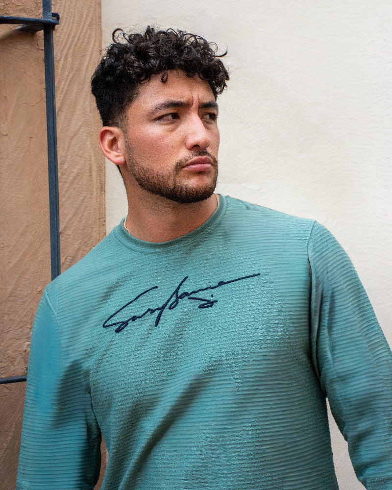 Hombre mirando hacia un lado con camiseta verde agua de textura lineal y gráfico de firma manuscrita en el pecho, marca Dubrovnik