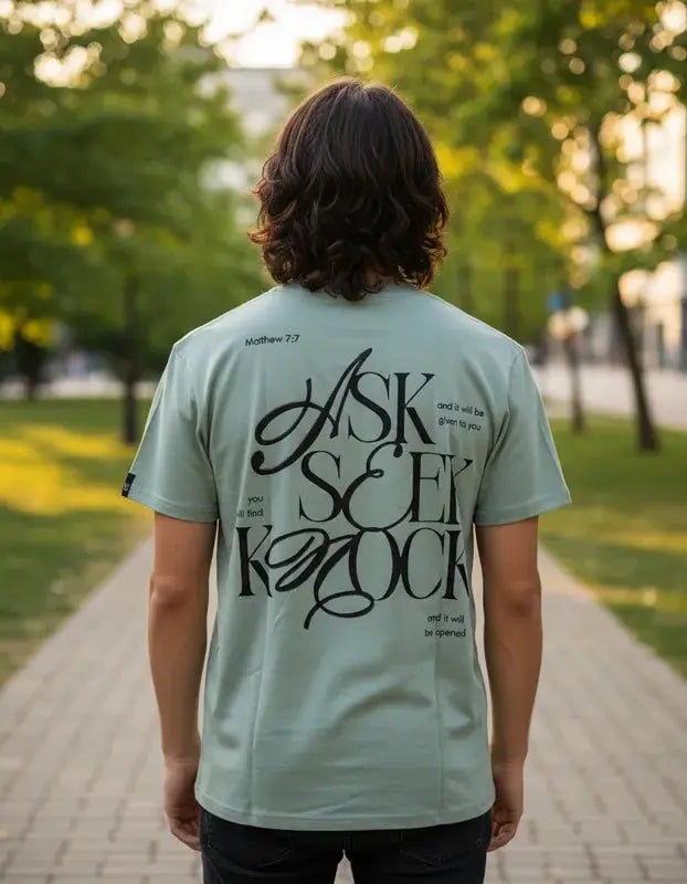 Vista trasera de hombre con camiseta color verde menta, destacando el diseño gráfico "ASK SEEK KNOCK" en color negro con la cita Matthew 7:7 en un parque