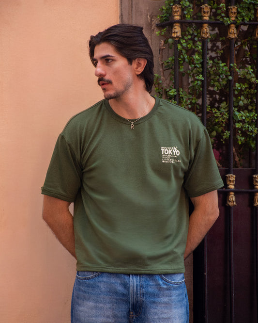 Hombre con bigote posando de perfil con camiseta verde oliva de manga corta, destacando un gráfico blanco con la palabra "TOKYO" y caracteres japoneses en el pecho.