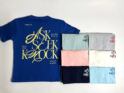 Camiseta azul royal extendida con diseño posterior amarillo "Ask Seek Knock" junto a una selección de camisetas dobladas en colores pastel, gris, blanco y marino de la marca Dubrovnik