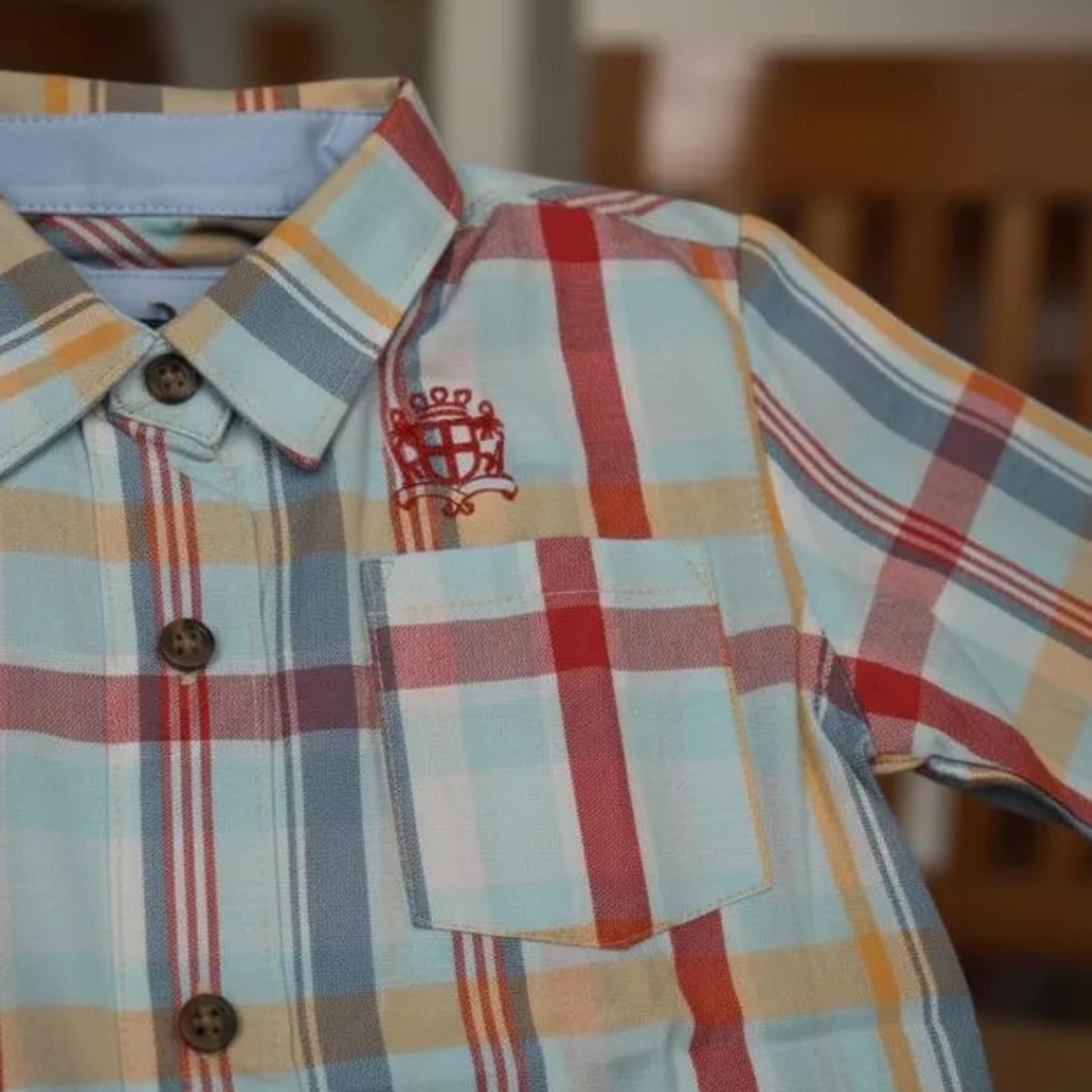 Primer plano de camisa infantil a cuadros celestes y naranjas con logotipo de corona bordado en rojo sobre el bolsillo