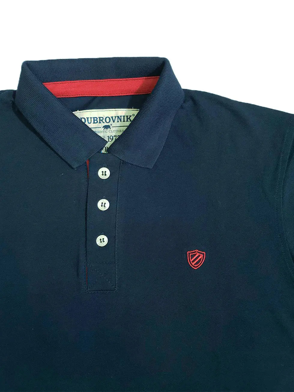Primer plano de un polo Dubrovnik azul marino mostrando el contraste granate en el cuello interno, botones reforzados y el logotipo del escudo bordado en rojo con precisión técnica