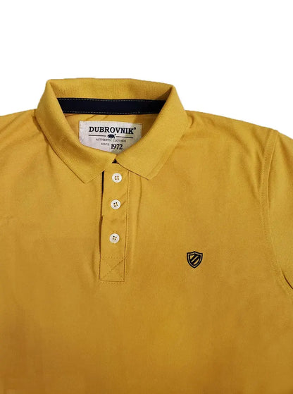 Primer plano de polo Dubrovnik color mostaza mostrando el contraste azul marino en el cuello interno, botones marfil reforzados y el escudo bordado con precisión técnica