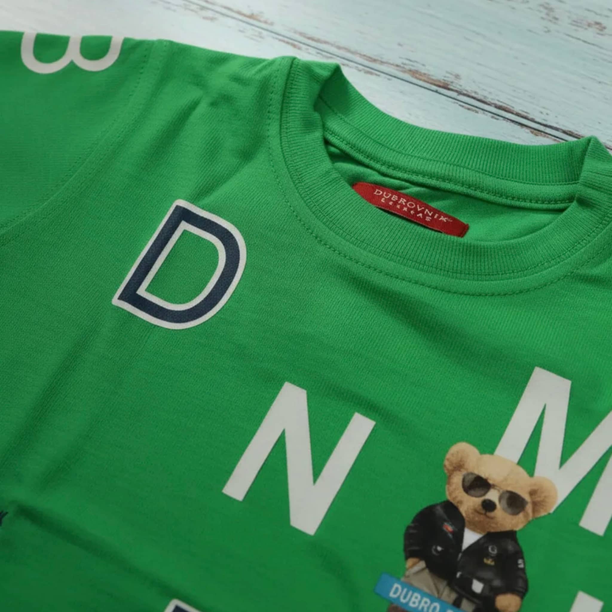 Primer plano del cuello de una camiseta verde para niño con etiqueta roja de Dubrovnik y estampado gráfico de letras blancas