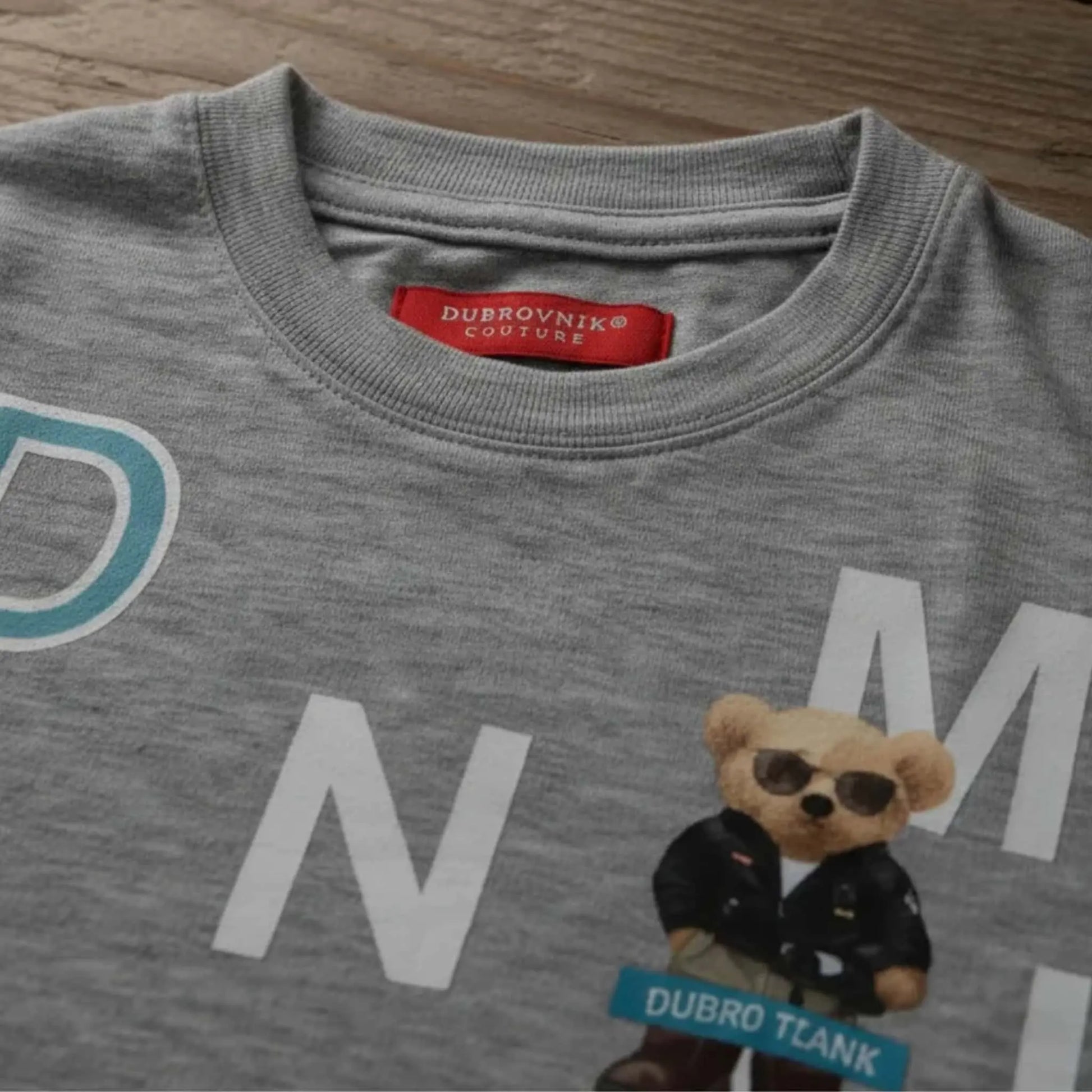 Primer plano del cuello de una camiseta gris jaspeada para niño con etiqueta roja "Dubrovnik Couture" y estampado de oso con gafas