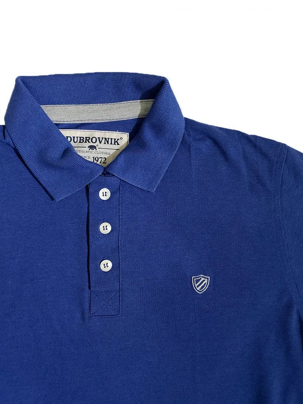 Primer plano del cuello de un polo infantil azul marino de la marca Dubrovnik, mostrando el ribete interior en gris, la etiqueta "Since 1972", tres botones blancos y el logotipo de escudo bordado en gris claro en el pecho
