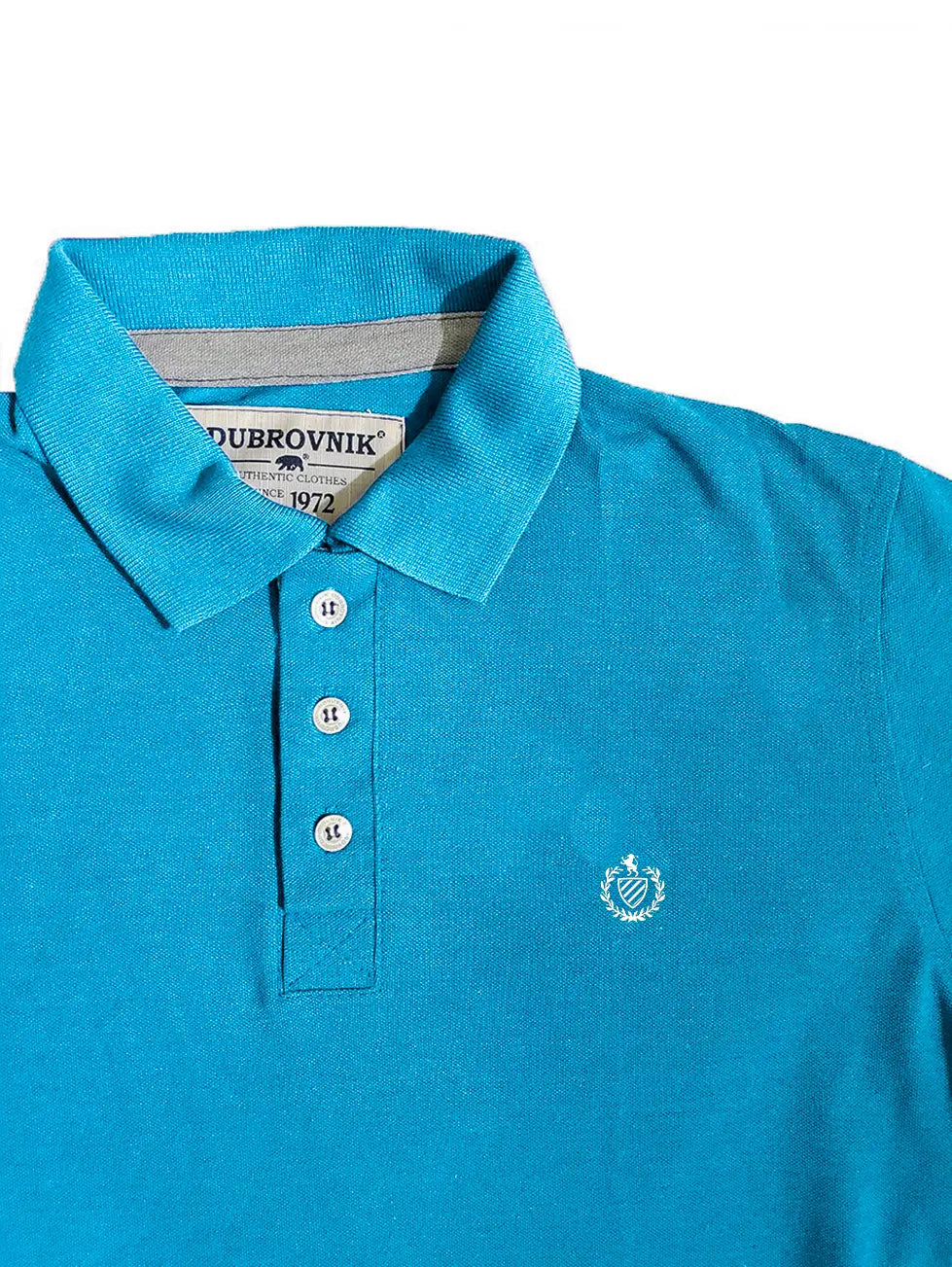 Primer plano del cuello de un polo infantil azul real de la marca Dubrovnik, mostrando el contraste gris en la tira interna del cuello, tres botones blancos y el logotipo del escudo bordado en blanco sobre el pecho.