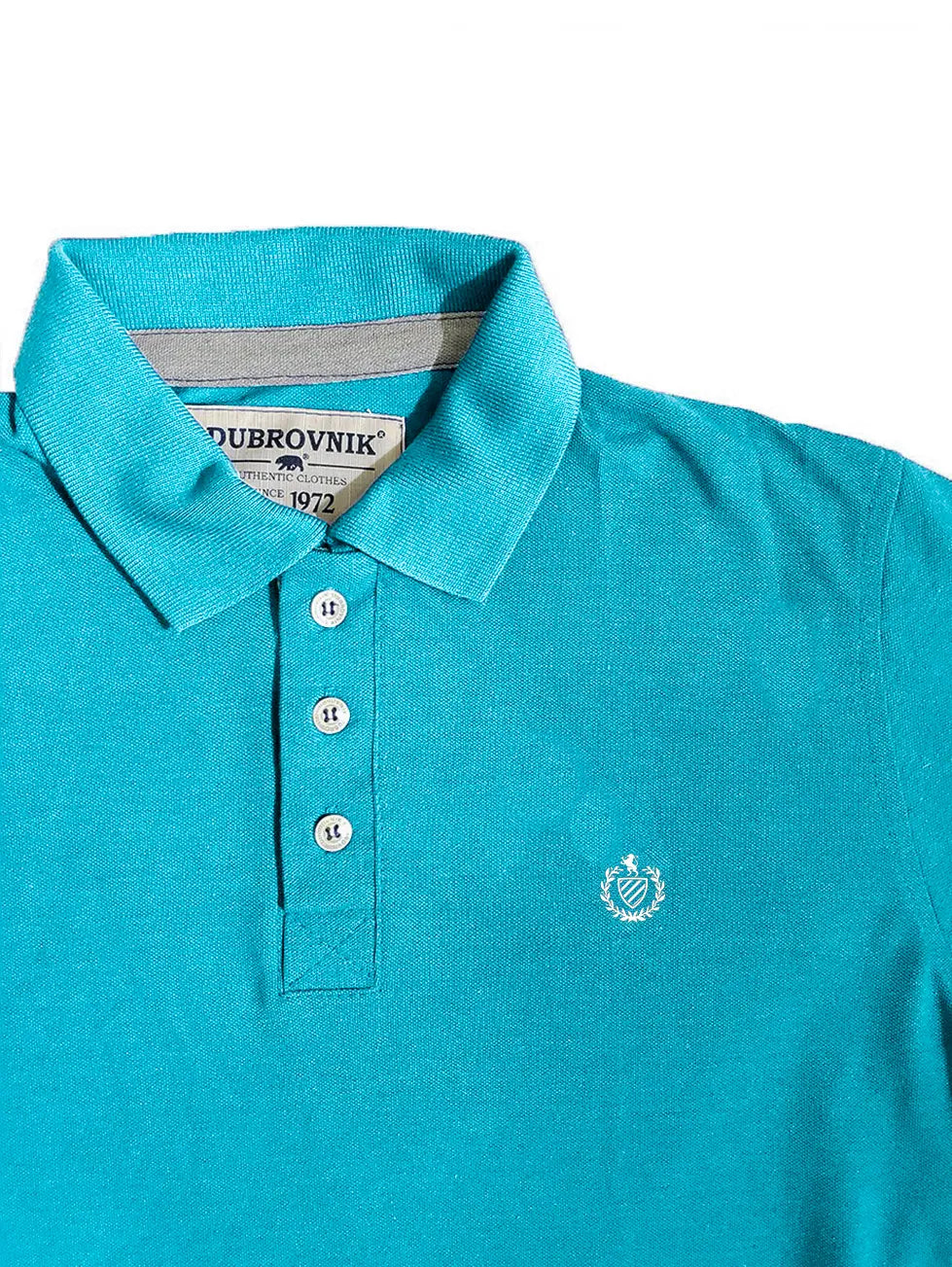 Primer plano real de un polo Dubrovnik color azul turquesa, mostrando el contraste interno en gris claro, botones color marfil y el escudo bordado en blanco con alta definición