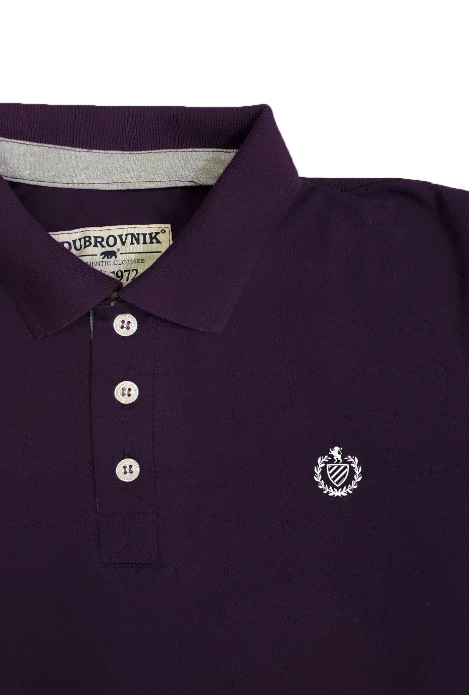 Primer plano real de un polo Dubrovnik color morado imperial profundo, mostrando el contraste interno en gris claro, botones perlados con cuatro orificios y el escudo bordado en hilo blanco de alta densidad