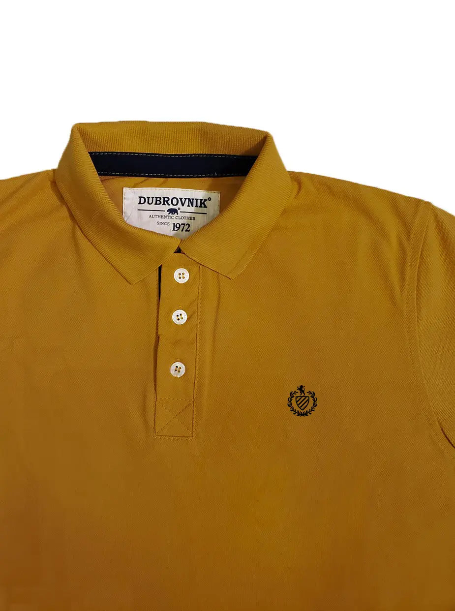 Primer plano real de un polo Dubrovnik color mostaza, mostrando la etiqueta tejida 'Authentic Clothes since 1972', el contraste azul marino en el cuello, los tres botones frontales y el escudo bordado en el pecho.