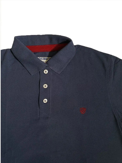 Primer plano de polo Dubrovnik azul petróleo, resaltando la cinta interna en color vino, botones reforzados y el escudo bordado a tono en el pecho