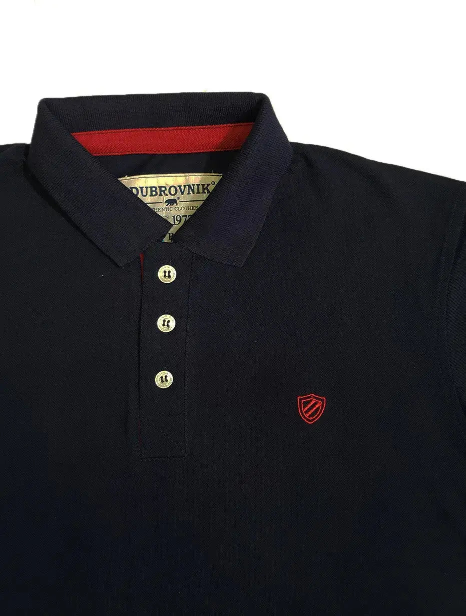 Primer plano de polo Dubrovnik negro, resaltando la cinta interna en color rojo vibrante, botones perlados y el escudo bordado circular en contraste