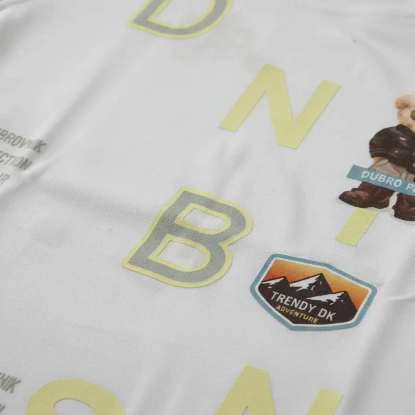 Primer plano de camiseta blanca con parches de montaña "Trendy DK Adventure" y estampado de letras amarillas de alta calidad