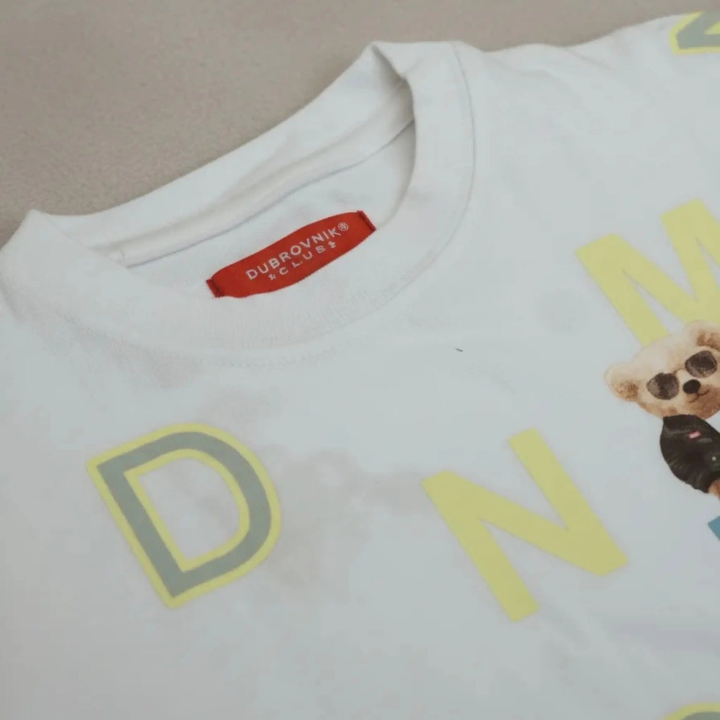 Primer plano del cuello de una camiseta blanca con etiqueta roja de la marca Dubrovnik Club y gráficos amarillos