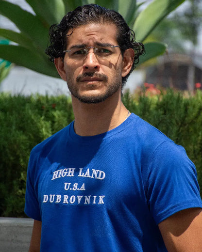 Primer plano de hombre con camiseta azul royal de manga corta destacando el bordado blanco de alta calidad "HIGH LAND U.S.A DUBROVNIK