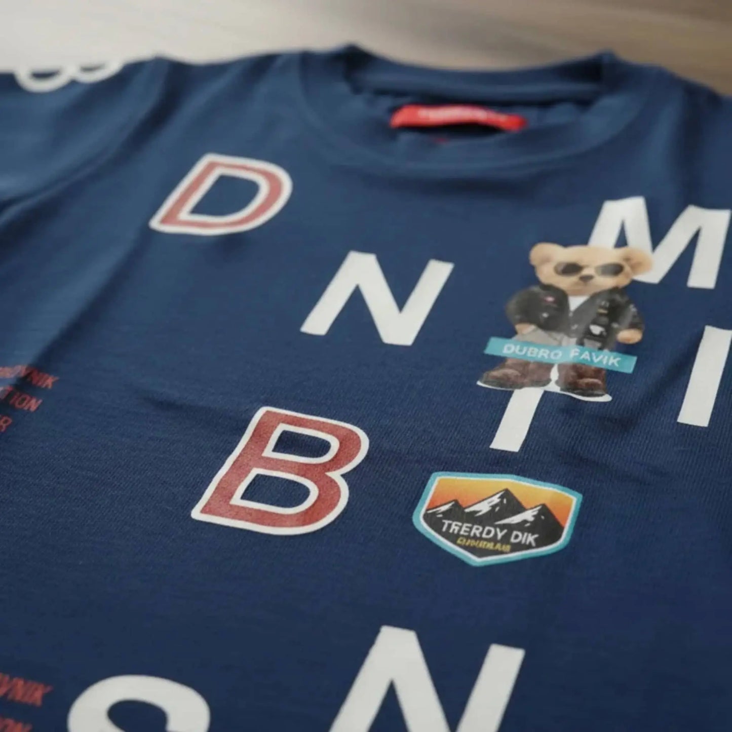 Primer plano de camiseta azul marino para niño con parches de oso con gafas, montañas "Trendy DK" y letras blancas y rojas