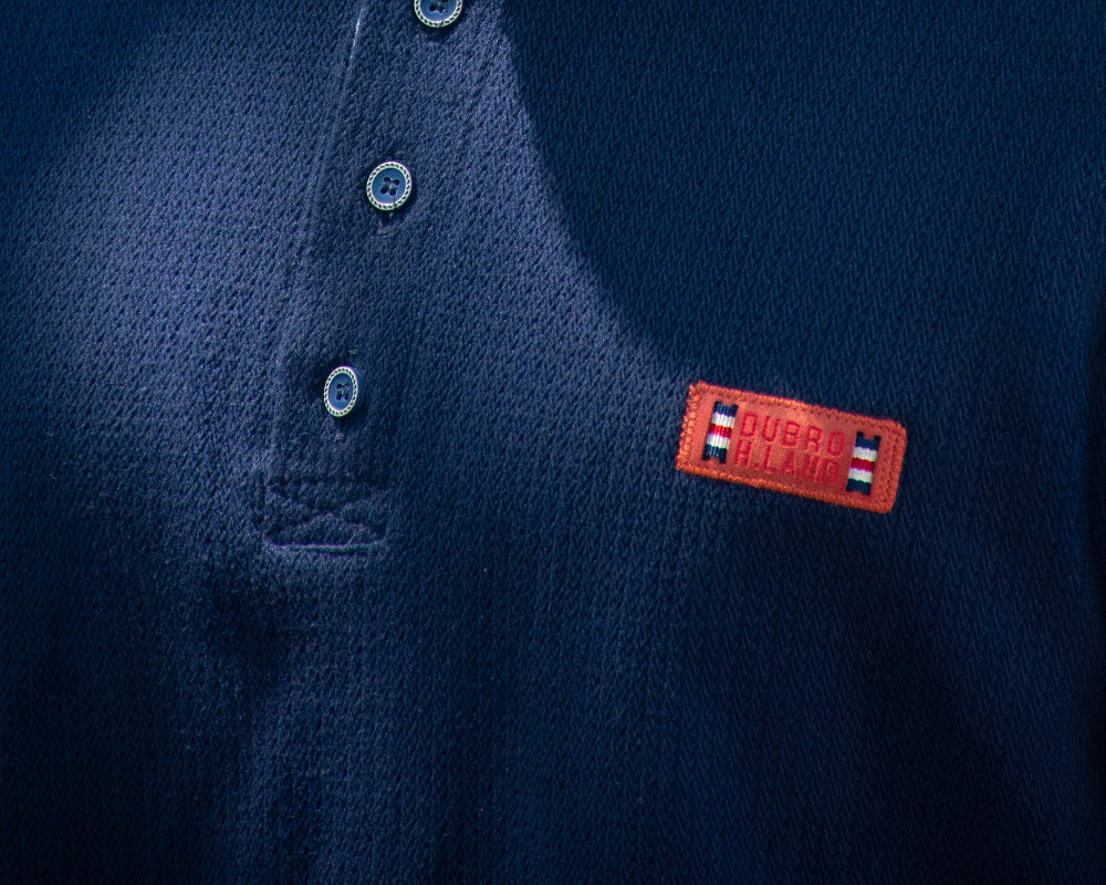 Primer plano del tejido texturizado azul marino de una camiseta Henley con botones y parche naranja bordado Dubro H.Land