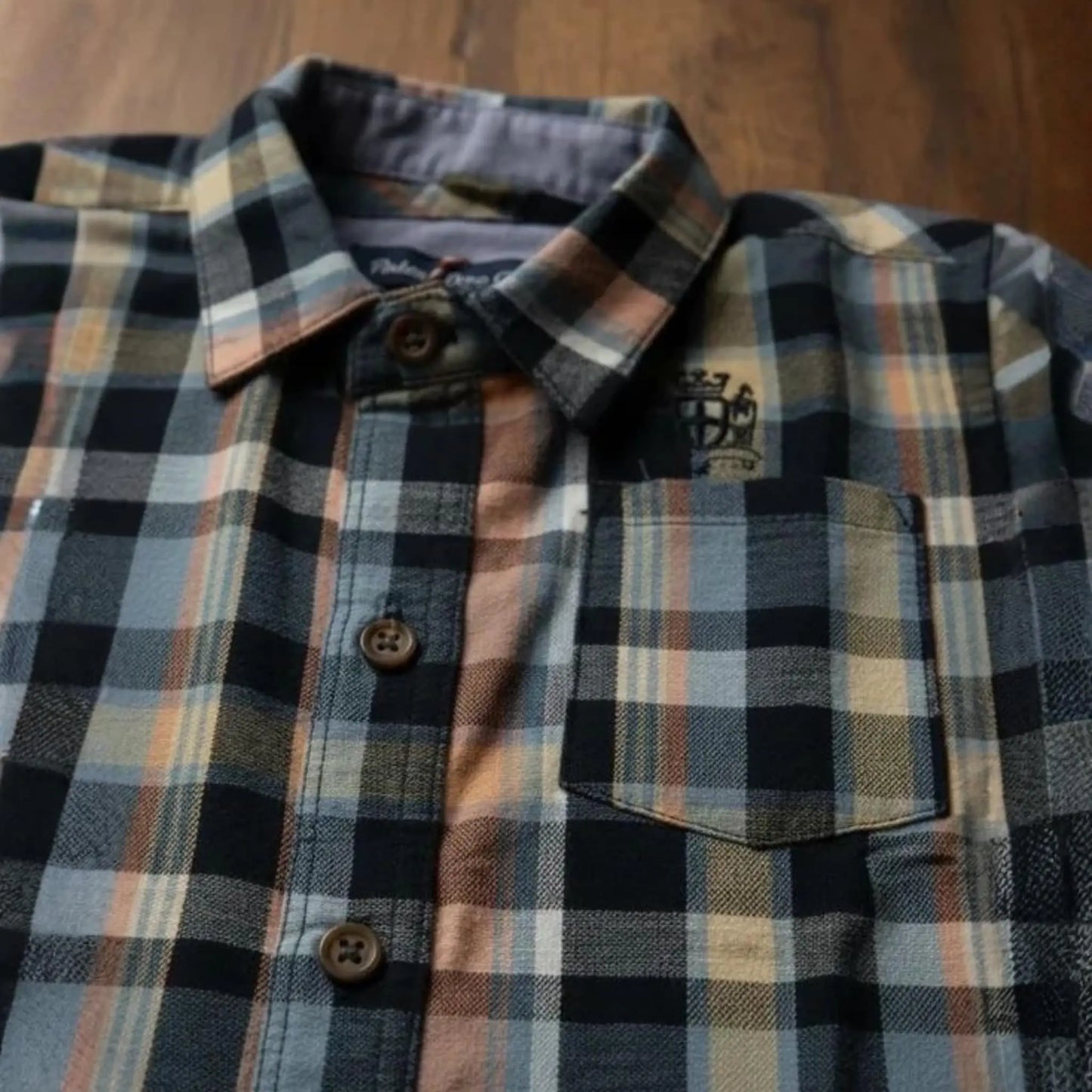 Primer plano de camisa infantil de cuadros con detalle de bolsillo frontal, botones de madera y logotipo bordado