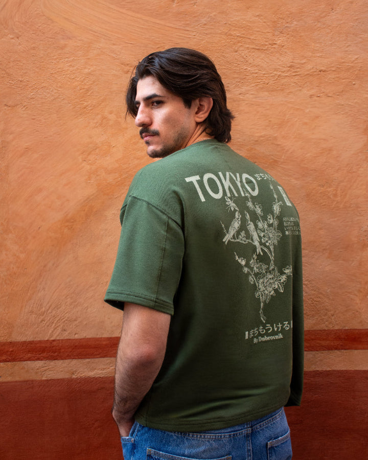 Plano posterior de una camiseta verde militar con un gran gráfico blanco que incluye pájaros en ramas, flores, caracteres japoneses y el texto 