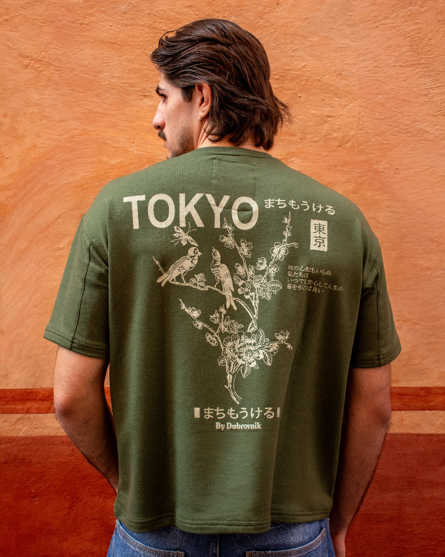 Vista trasera de camiseta verde militar con ilustración blanca de pájaros en ramas, letras japonesas y el texto TOKYO By Dubrovnik