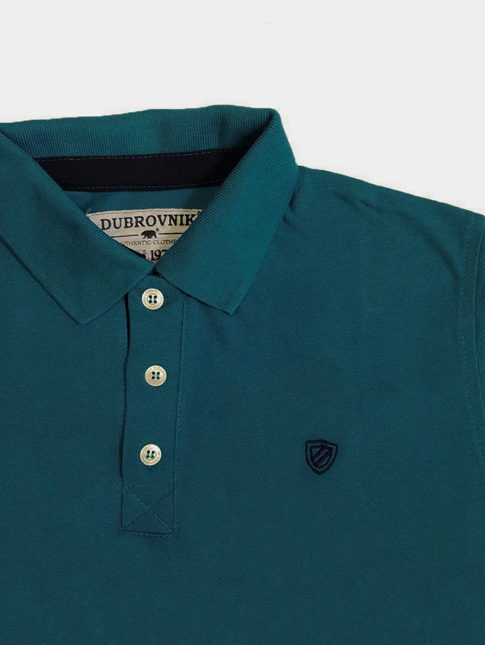 Primer plano del cuello de un polo infantil en color azul petróleo de la marca Dubrovnik, mostrando el contraste marino en la cinta interna, botones claros con acabado brillante y el escudo bordado a tono en el pecho.