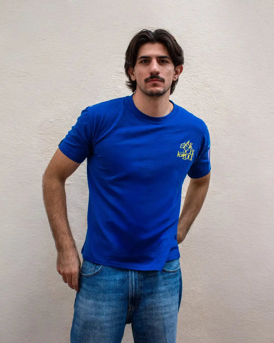 Hombre con bigote posando de frente con camiseta azul royal, mostrando el logotipo "Ask Seek Knock" en pequeño en el pecho y vaqueros denim.