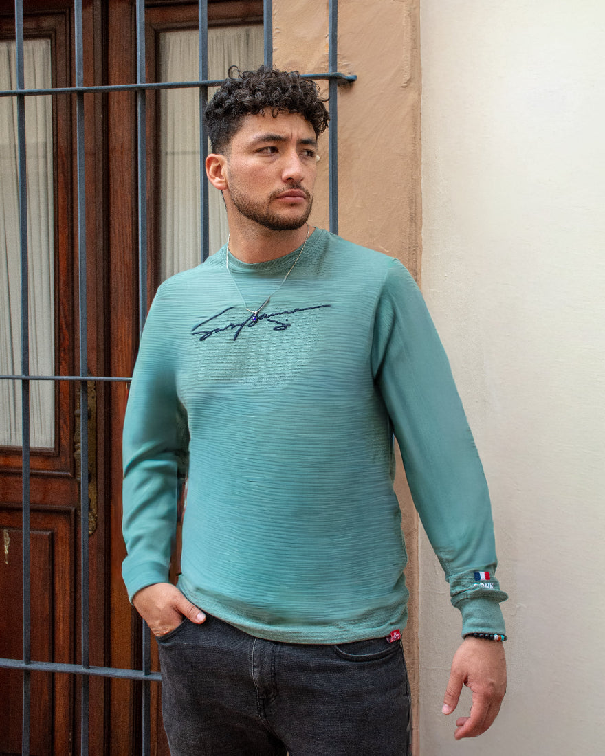 Hombre con jersey de manga larga verde agua texturizado, mostrando detalle de bandera en el puño y gráfico de firma en el pecho