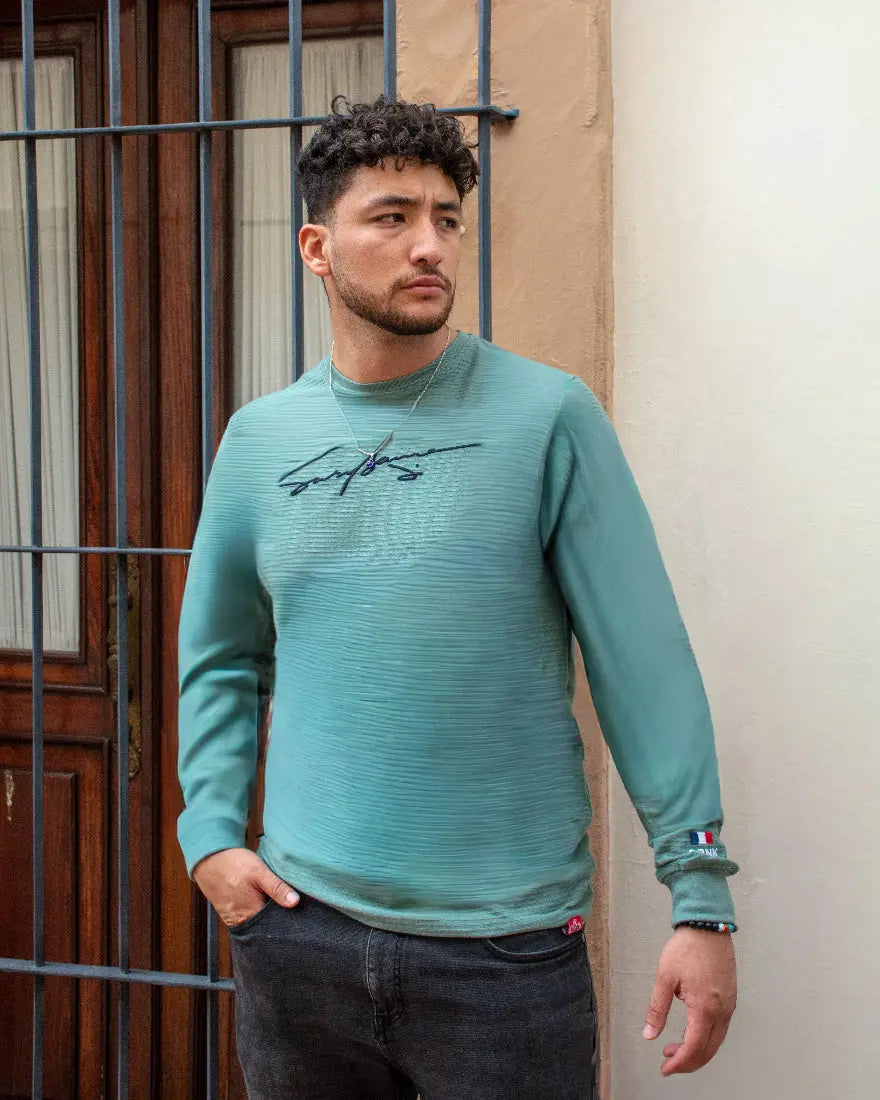 Hombre con jersey de manga larga verde agua texturizado, mostrando detalle de bandera en el puño y gráfico de firma en el pecho
