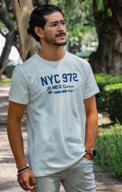 Hombre con gafas y reloj apoyado en un árbol, vistiendo una camiseta blanca con estampado gráfico azul "NYC 972" en un entorno de parque natural
