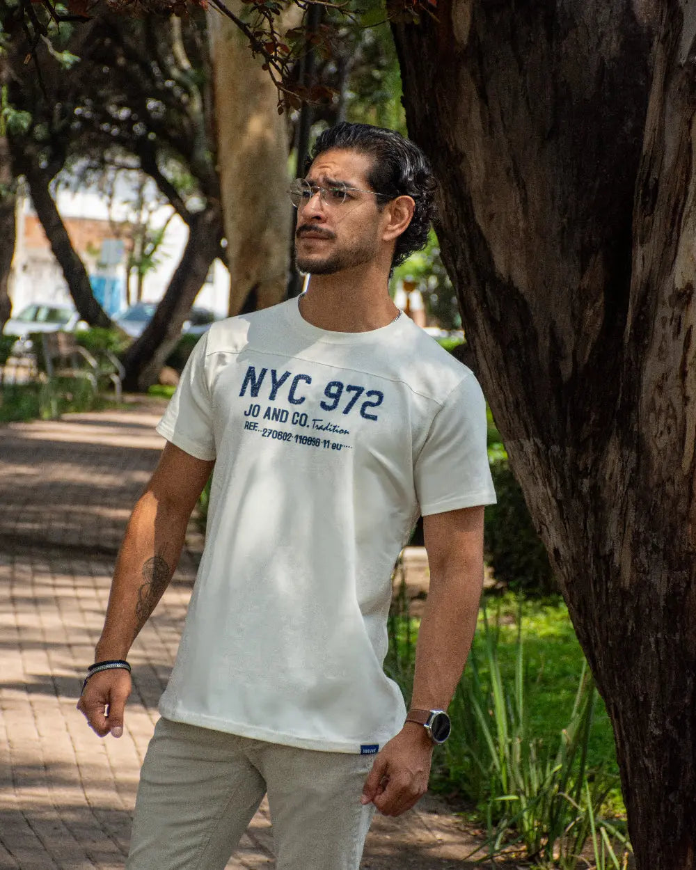 Hombre apoyado en un árbol vistiendo camiseta blanca con gráfico azul NYC 972, gafas de ver y reloj, en un parque soleado